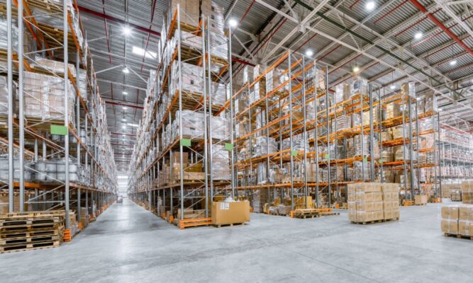 A large warehouse lit with LED high bay lights features tall metal shelving units stacked with cardboard boxes.
