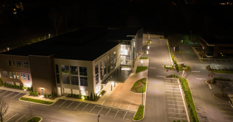 An aerial view of an office building at night features a well-lit parking lot and green landscaping.